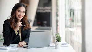 Alt image text:  A woman in business attire sitting at a table, holding a pen with her laptop open and smiling at the camera