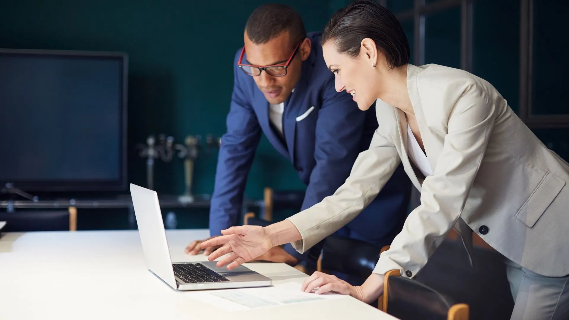 A woman dressed in business attire pointing to a laptop screen while explaining something to a male colleague also dressed in business attire. 