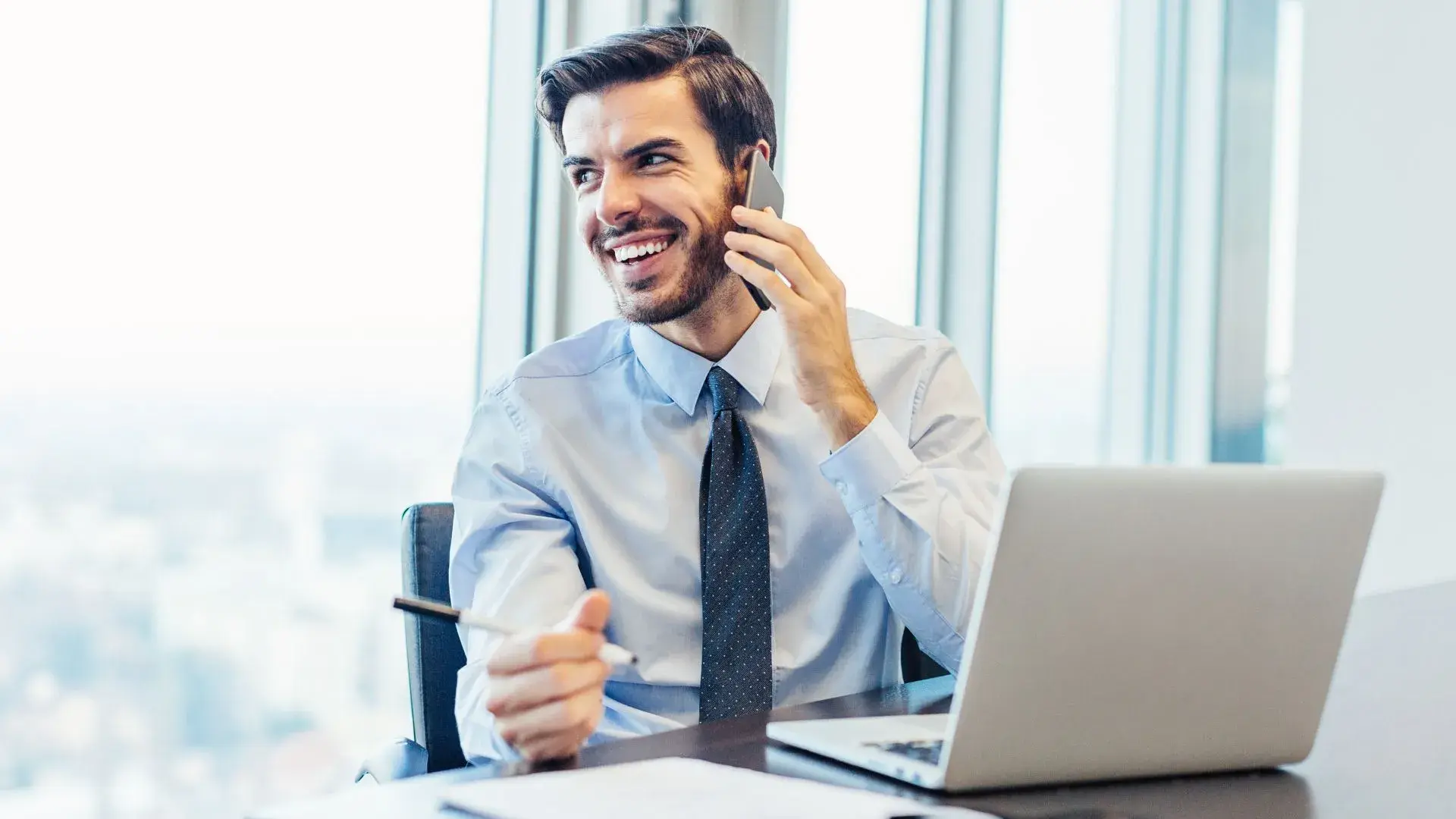 A man in corporate attire using his phone while working over his laptop