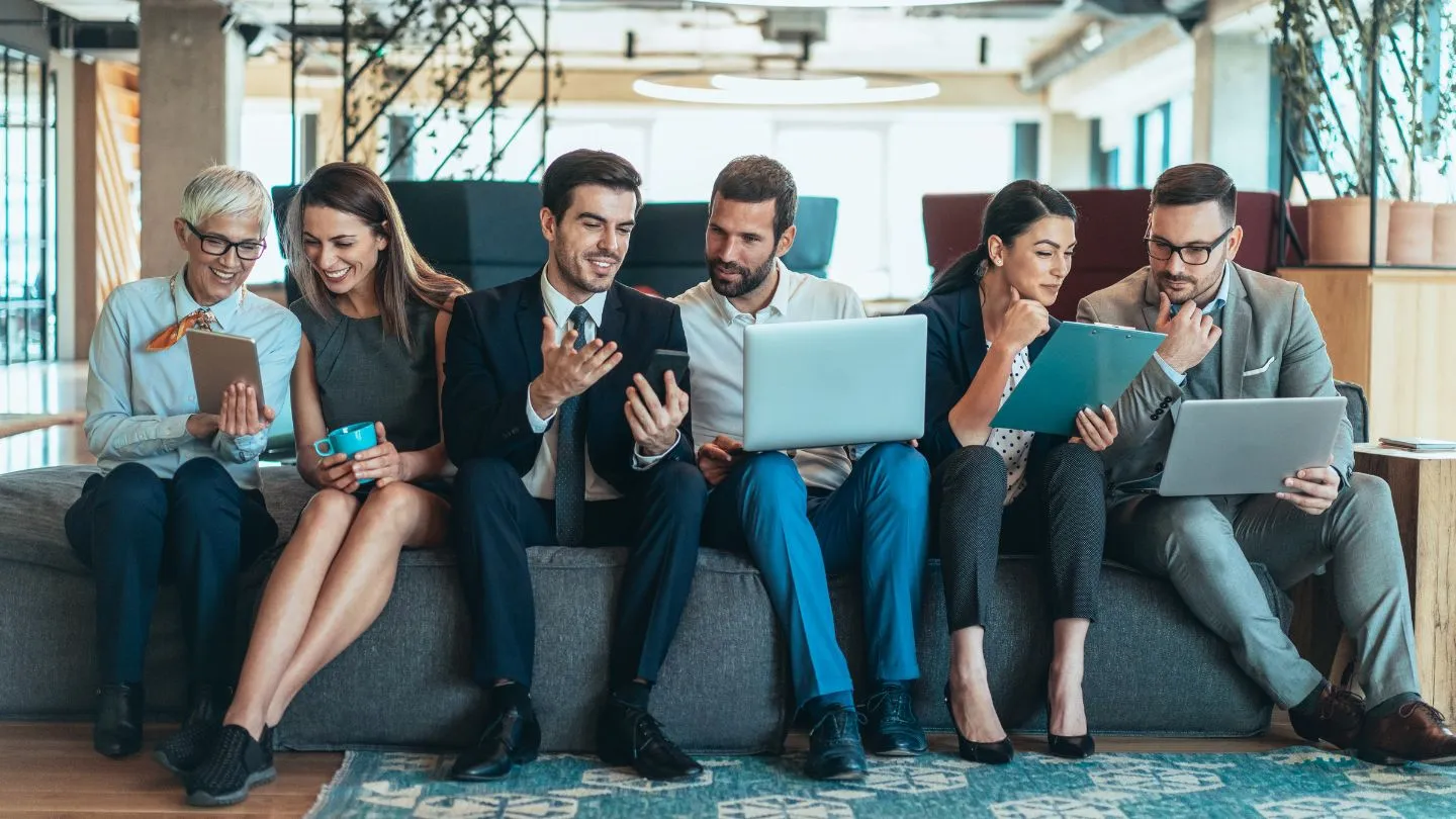 A group of office employees are seated on a couch, with small groups in intent discussions over laptops, phones, and tablets   